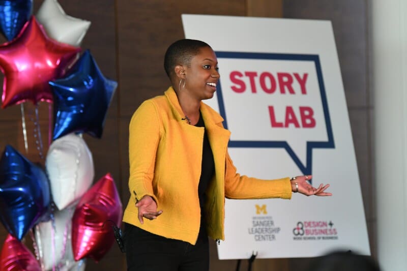 A speaker in a yellow blazer presenting at the Story Lab event, hosted by the Sanger Leadership Center and Ross School of Business Design + Business program, with star-shaped balloons visible in the background.