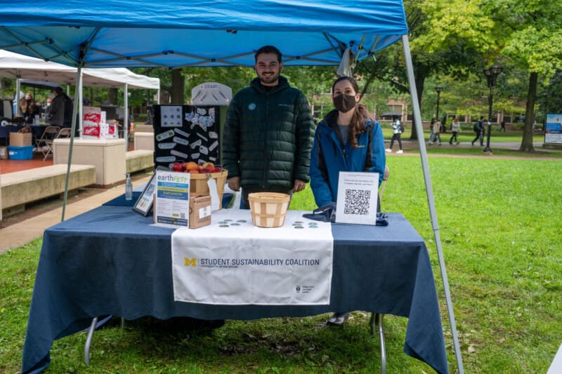 Two University of Michigan students staffing a Student Sustainability Coalition table at an outdoor campus event, with EarthFest materials, a basket of apples, and a QR code sign displayed on the table.