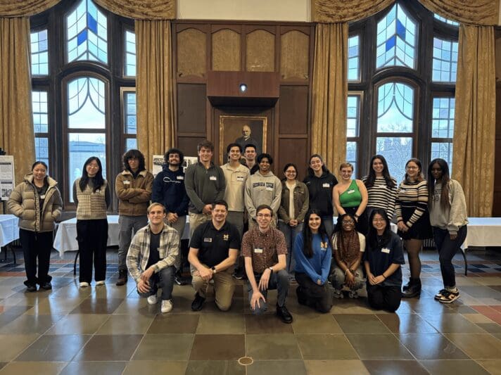 A group of University of Michigan students and facilitators posing together inside an elegant wood-paneled room with tall arched windows, wearing name tags at an organizational leadership program event.