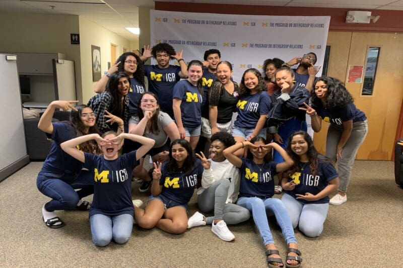 A large group of University of Michigan students posing and smiling together, many wearing navy blue IGR t-shirts, in front of a "Program on Intergroup Relations" branded backdrop.