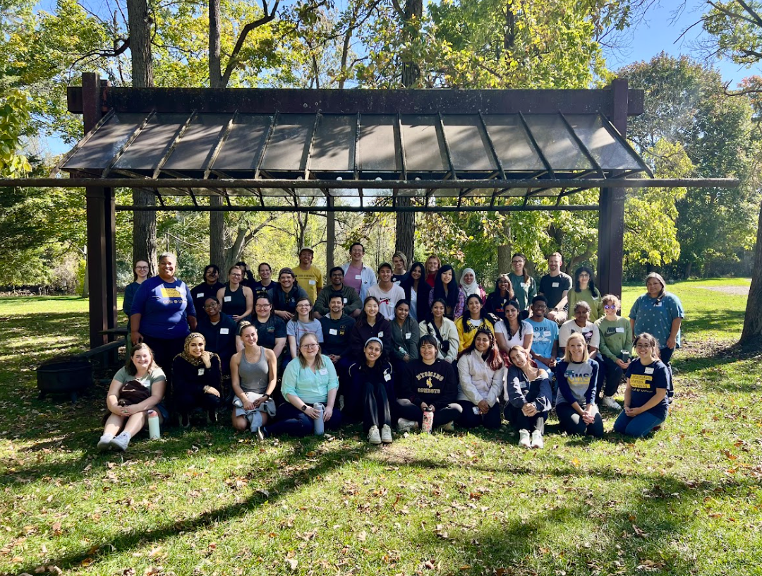A large group of students and facilitators posing together outdoors under a wooden pergola structure surrounded by autumn trees, wearing name tags at an inclusive leadership retreat.